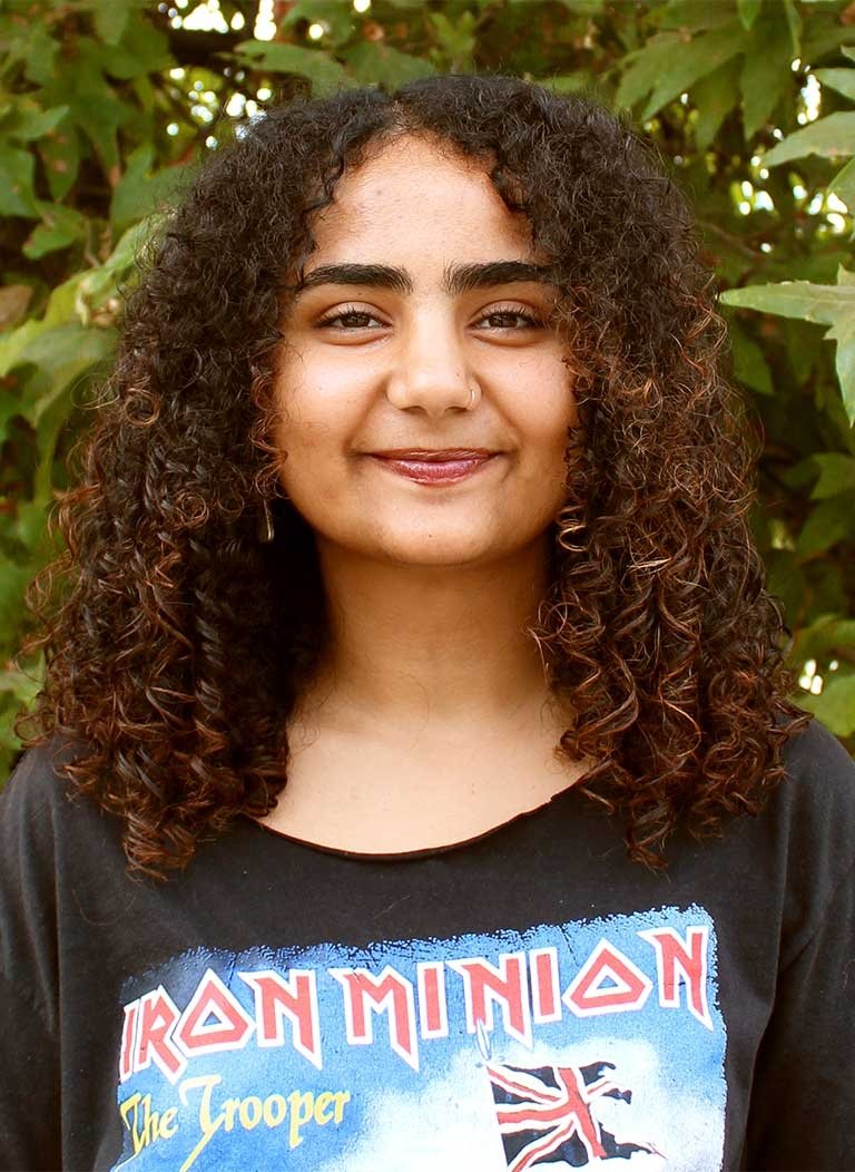 a student with curly long brown hair wears a iron minion tshirt while posing in front of a bush