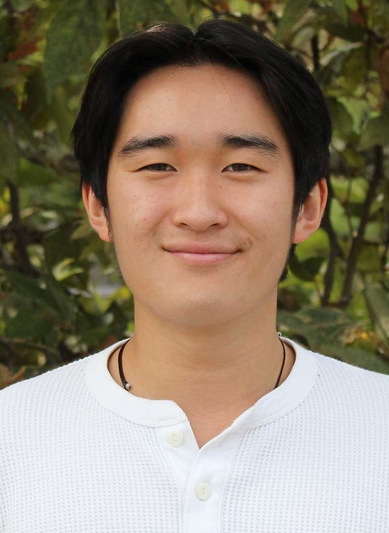 a student with short black hair wears a white henley shirt while standing in front of outdoor bushes