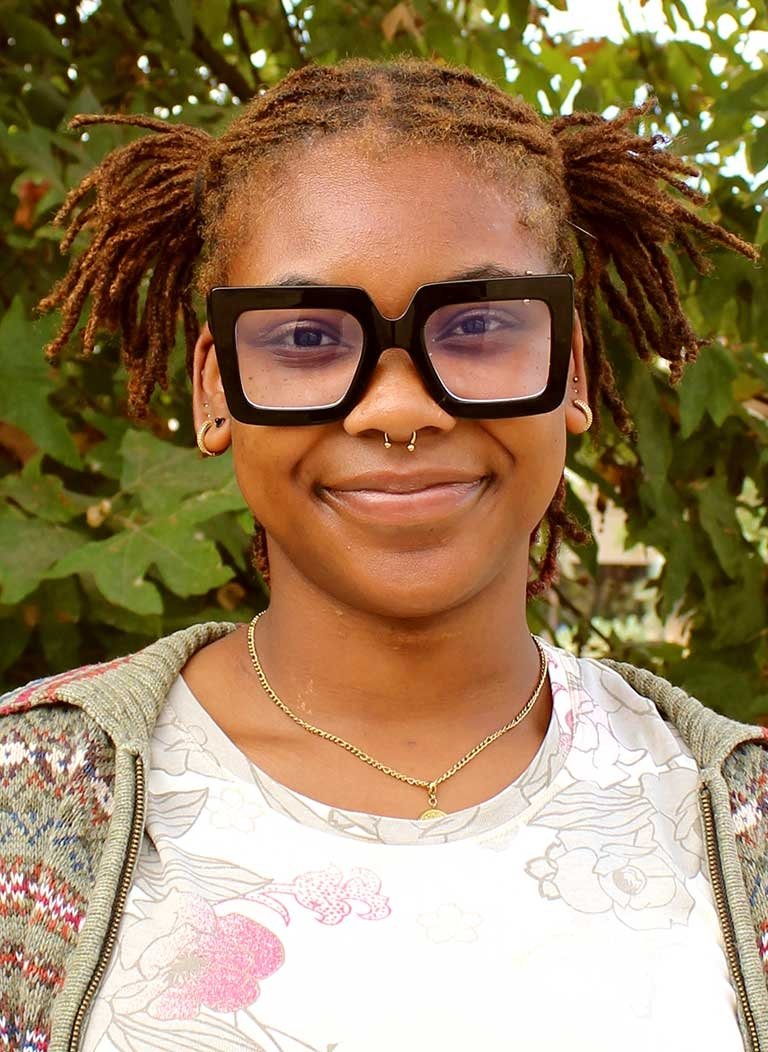 a student wearing a tshirt, sweater, and black glasses stands in front of outdoor landscaping