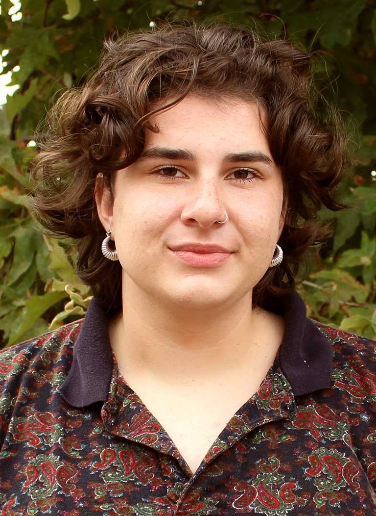 a student with short brown hair stands in front of outdoor greenery