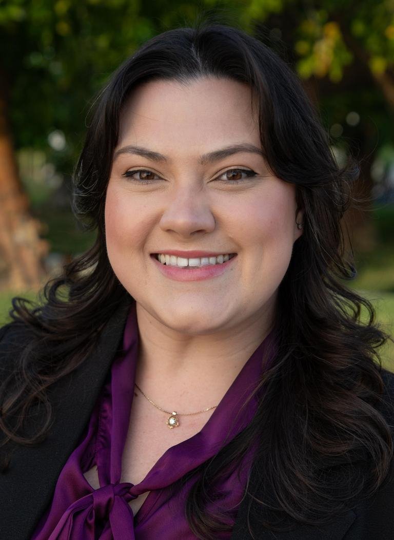 Woman in purple blouse wearing black jacket with curly black hair.