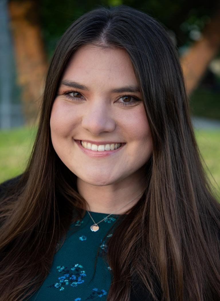 Woman in green floral top with black blazer and long brown hair. 