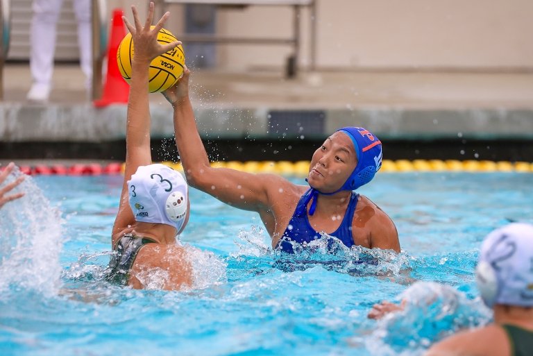 An action shot of water polo player Namlhun Jachung in the pool. Another player attempts to block as Jachung holds up the yellow ball.