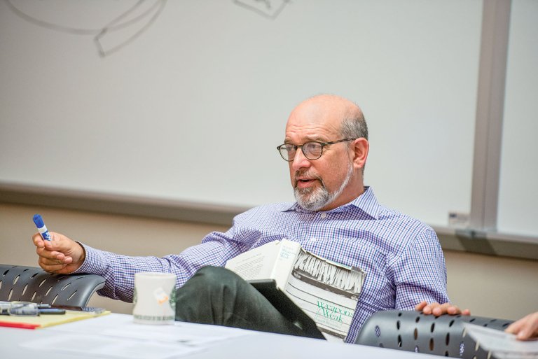 Dan Segal wearing glasses blue stripped shirt, sitting in a classroom