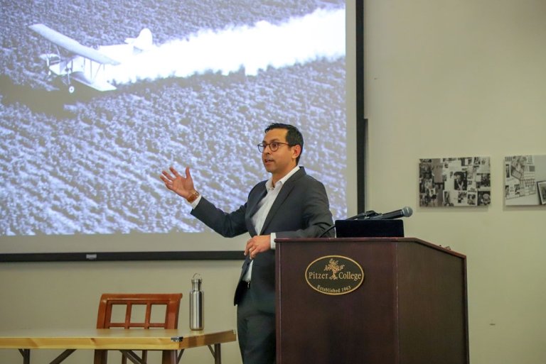 Adam M. Romero stands at the podium with the Pitzer College logo. A photo of a crop duster appears on the projector screen behind him.