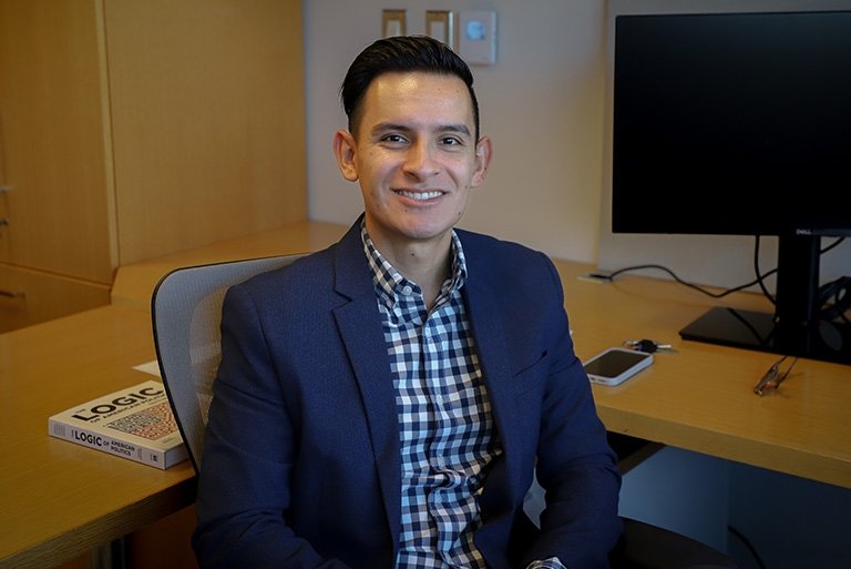 Cesar Vargas Nunez sits in his office. He has short dark hair and wears a navy blue blazer over a white and blue plaid shirt.