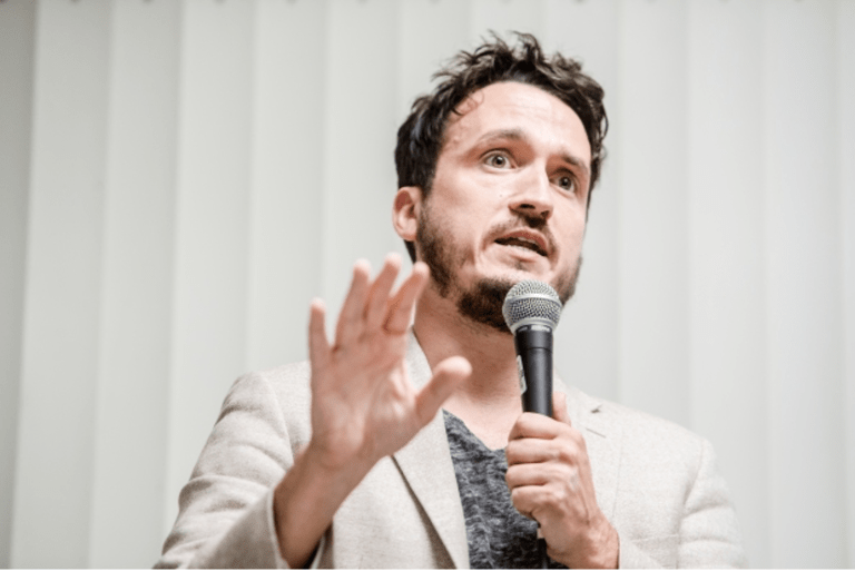 Michael Ceraso speaks on a microphone, wears a white blazer, and has short dark hair and beard.
