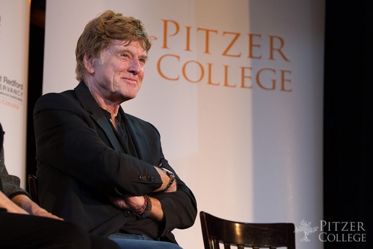 Robert Redford with short light brown hair and wearing a black blazer sits in front of a step-and-repeat with orange text that reads Pitzer College.