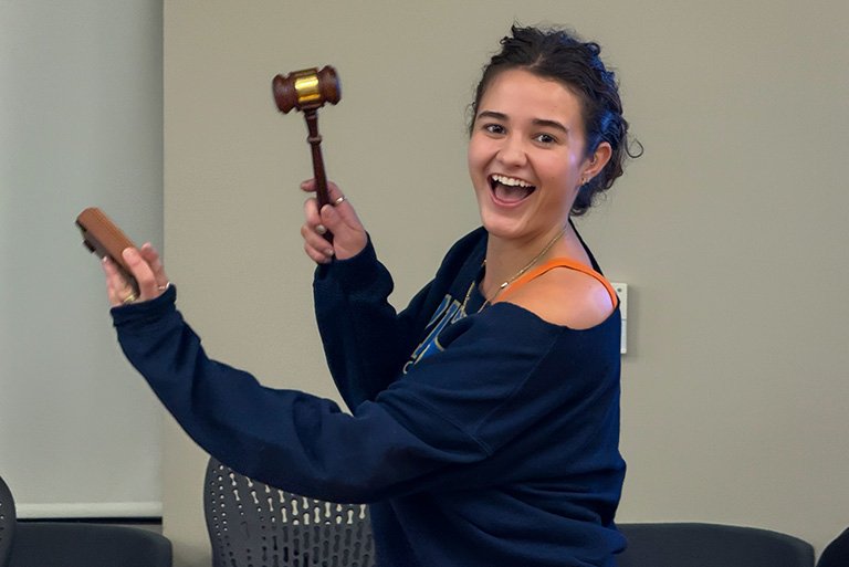 Ella Hale holds a gavel and grins while standing in a conference room