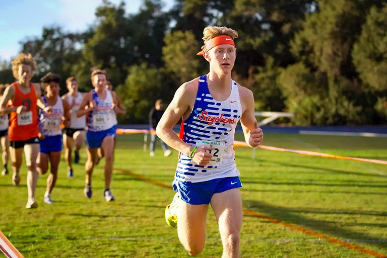 a student in a blue and white and orange tank top and blue shorts runs in a race with competitors in the background