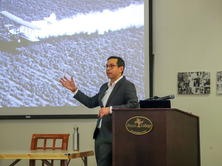 Adam M. Romero stands at the podium with the Pitzer College logo. A photo of a crop duster appears on the projector screen behind him.