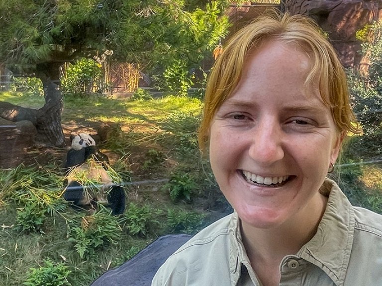 Jana Biedenweg stands in front of the giant panda enclosure at San Diego Zoo while a panda eats bamboo.