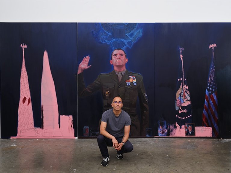 Vincent Valdez crouches in front of a large painting. Valdez is bald and wears glasses, a gray T-shirt and dark pants.