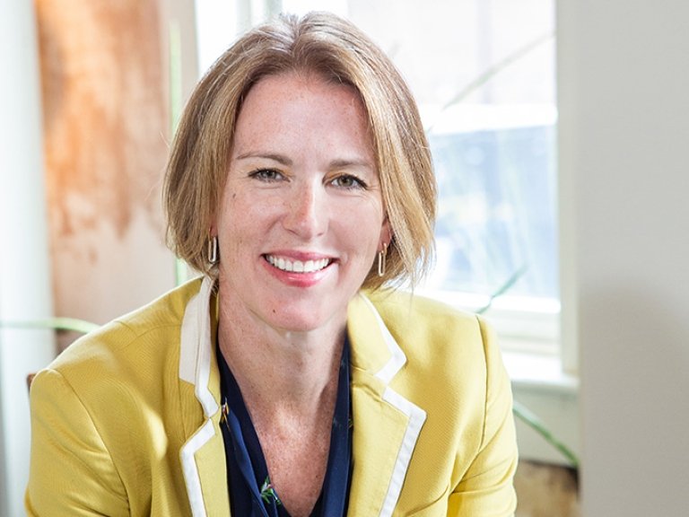 Tricia Milford-Hoyt has straight blond hair in a bob cut and wears a yellow blazer over a blue shirt.