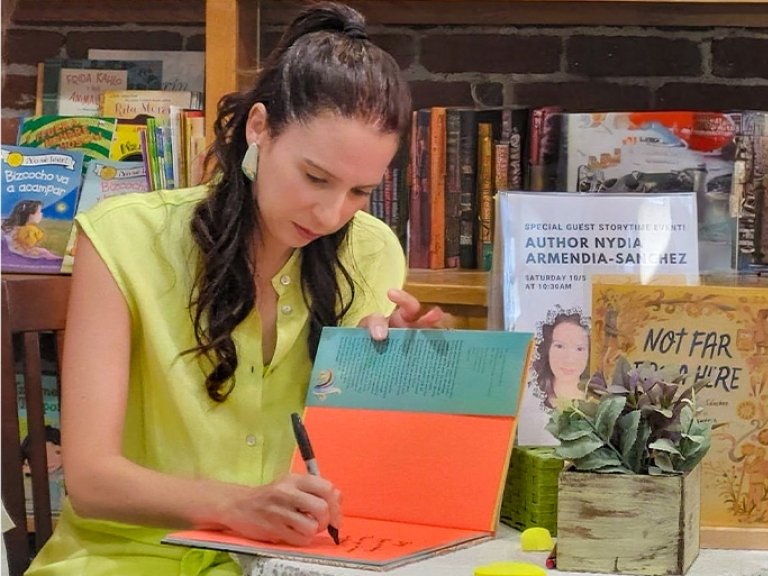 Nydia Armendia-Sanchez has long dark hair and wears a pale yellow top while signing a children’s book.