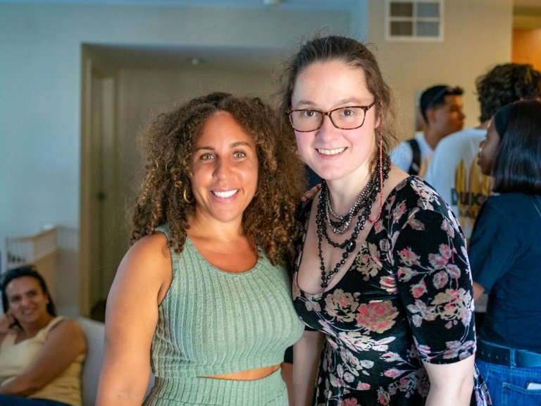 Professor Fely Catan wears teal outfit and Professor Amanda Louise Johnson wears a floral dress inside a faculty apartment.