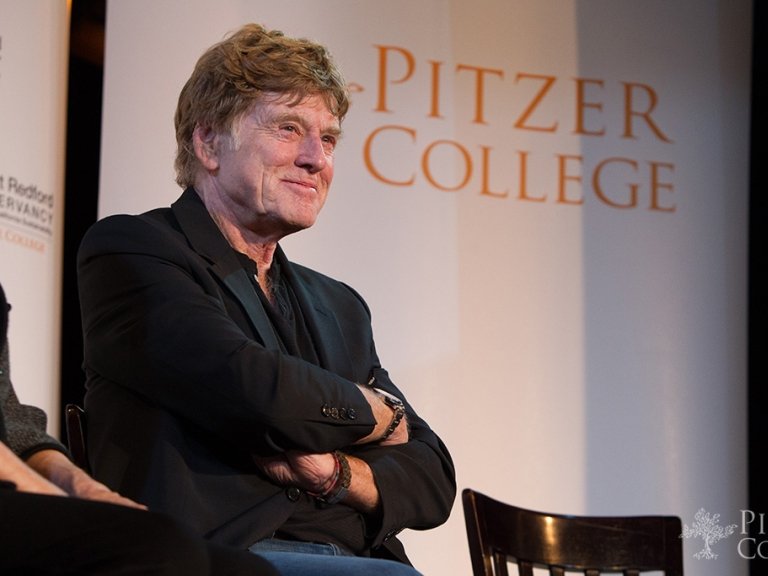 Robert Redford with short light brown hair and wearing a black blazer sits in front of a step-and-repeat with orange text that reads Pitzer College.