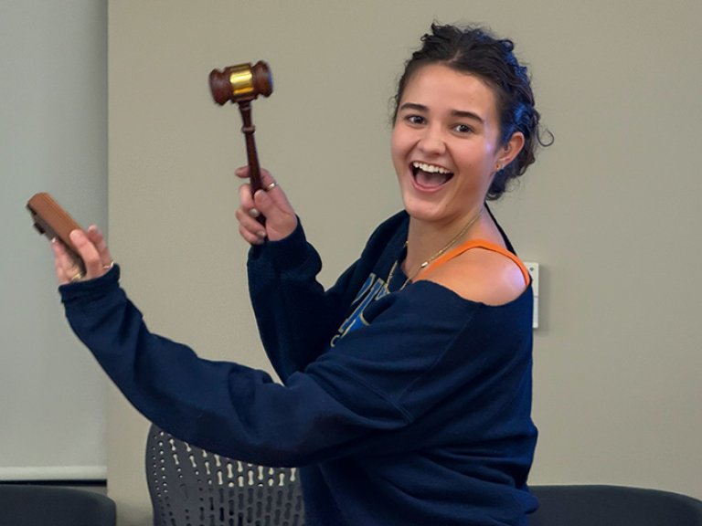 Ella Hale holds a gavel and grins while standing in a conference room