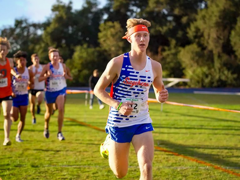a student in a blue and white and orange tank top and blue shorts runs in a race with competitors in the background