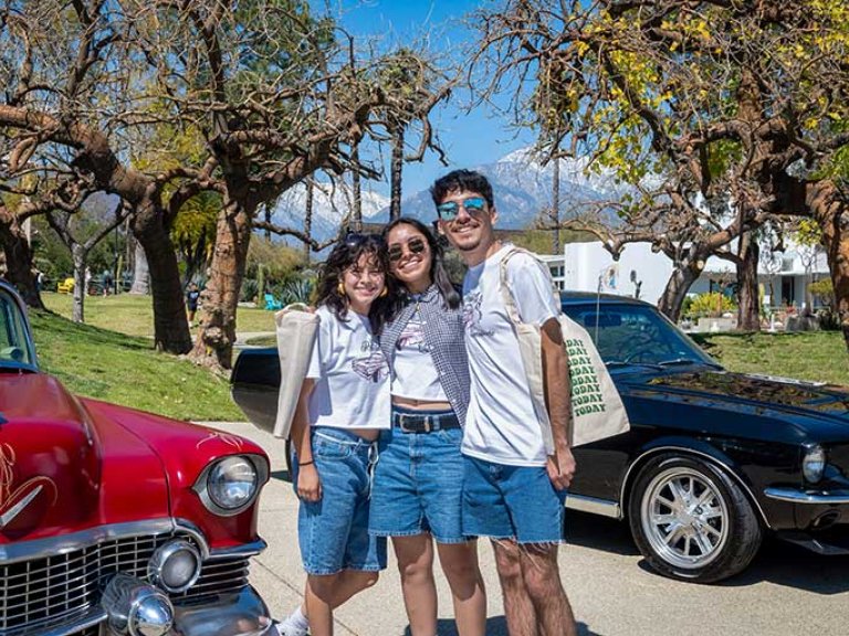 three students pose in front of two classic cars parked near the mounds