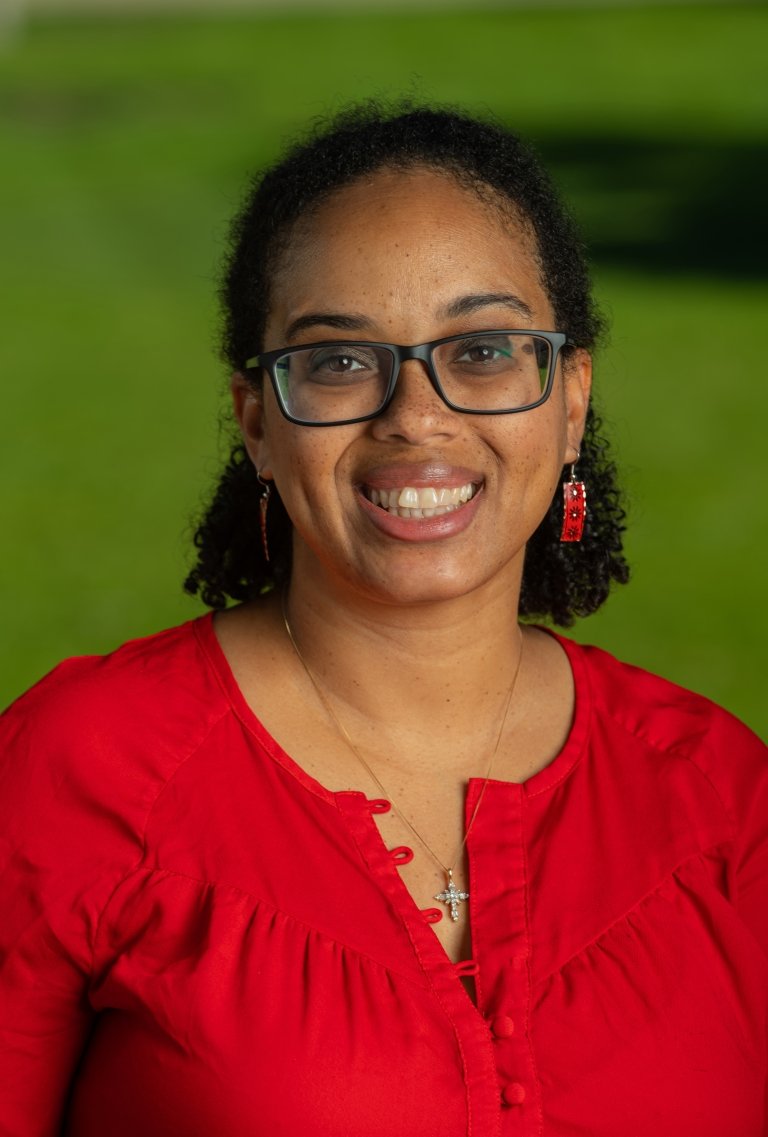 Woman wearing glasses and wearing red earrings and red blouse, standing in front of greenery.