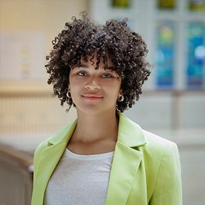 Close-up of Corrine Waters, who has curly brown hair and wears a pale green blazer over a white shirt.