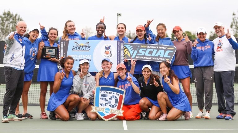 The Pomona-Pitzer women’s tennis team gathers on the court for a group photo. The team raises their fingers in a number one gesture and hold up signs for the SCIAC tournament championship.