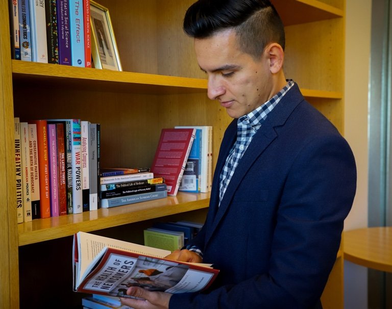 Cesar Vargas Nunez stands in front of his bookshelf as he peruses a book.