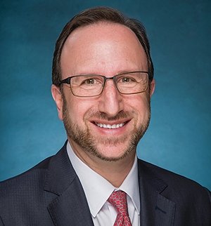Man with short brown hair wearing glasses and grey suit with red tie.