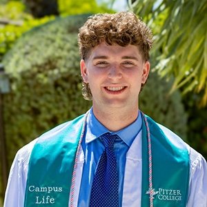 Close-up of Jack Pine with short curly brown hair and wearing a teal stole and white graduation robe over a blue collared shirt and tie.