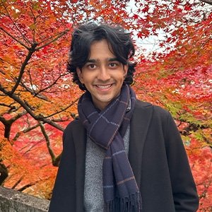Close-up of Tommy Shenoi with medium-length black hair and wearing a black coat, gray sweater, and dark blue scarf.