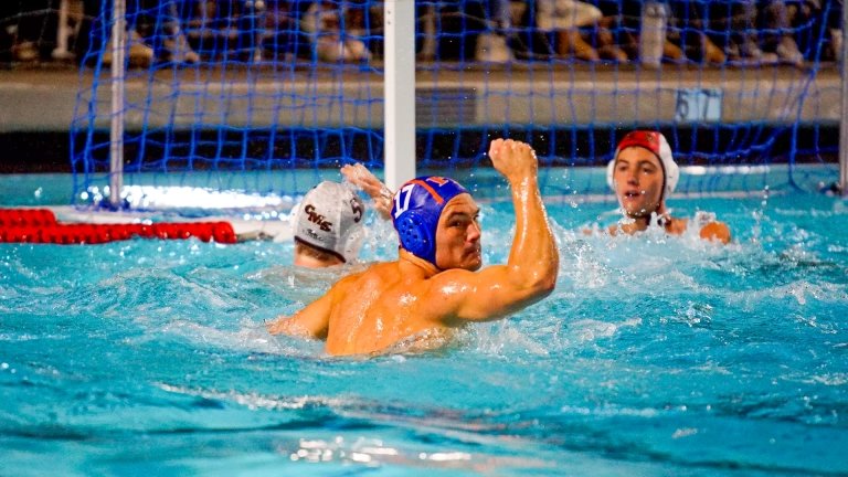 A member of the Pomona-Pitzer men’s water polo team wearing a blue swim cap holds up a fist victoriously in the pool.