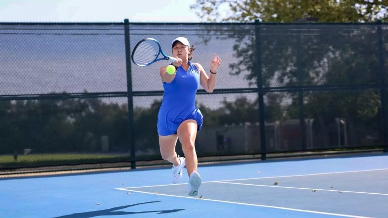 A Pomona-Pitzer women’s tennis player wearing a blue uniform swings a racket on the court.