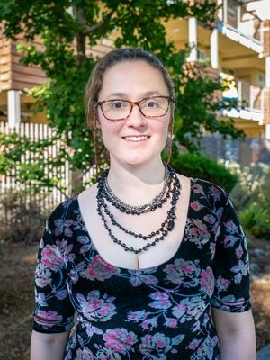 Amanda Louise Johnson has brown hair pulled back and wears glasses, a floral top, and beaded necklaces.