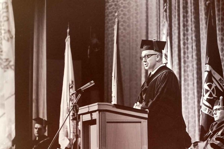 john atherton wears a cap and robe while speaking a podium on a stage 