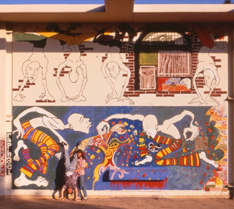 aaron rhodes does a handstand next to Darci Raphael in front of a colorfule mural of dancing human figures