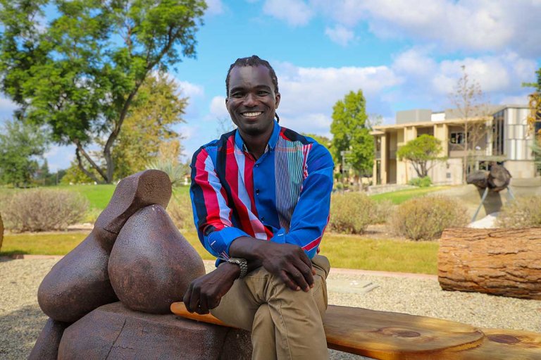 Batsirai Chidzodzo sites on a bench in Holden Garden with the Mound and Avery behind him