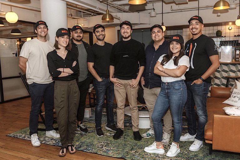 a group of several people stand in a loft wearing baseball caps that say 'maza'