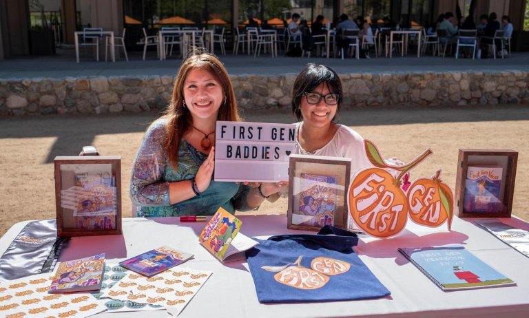 two pitzer student staff sit a the first gen table holding up a sign that says "first gen baddies"