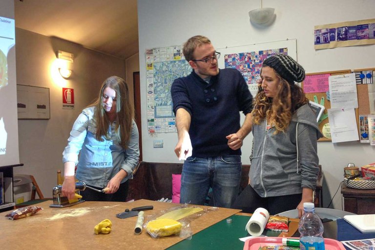 one student makes pasta while another listens to the instructor while standing in a classroom