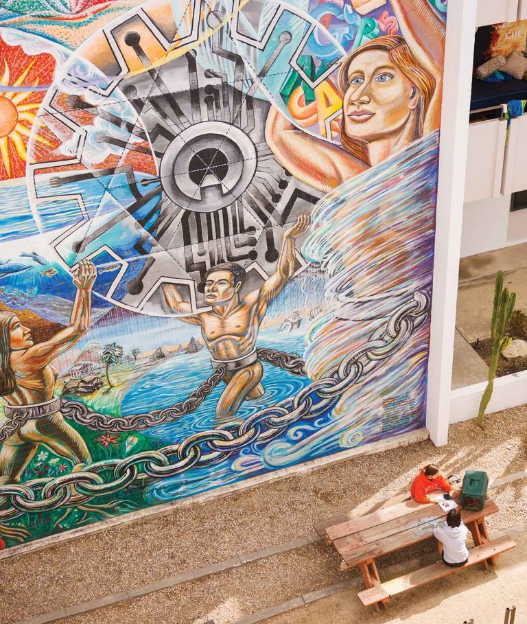 two students sit at a picnic bench with a large colorful mural behind them