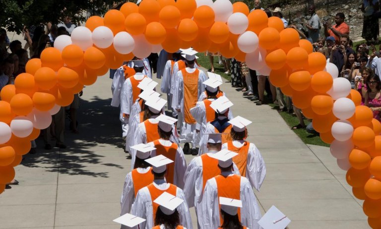 graduates in full regalia walk under an orange and white balloon arch