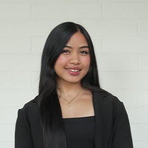 Janae Garcia has long black hair and wears a black top while standing against a white wall.