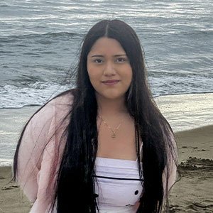 Leslie Valois has long dark hair and wears a pale pink tope while standing by the ocean.