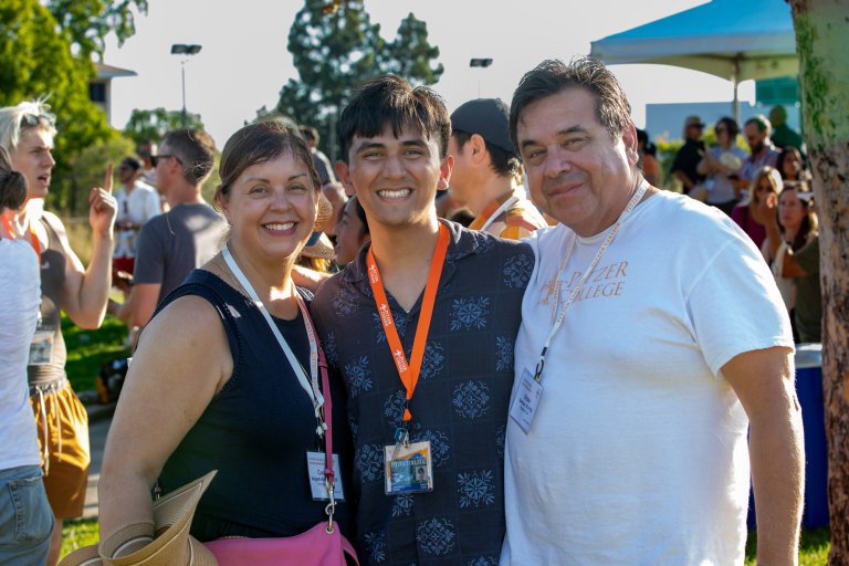 A Pitzer student and their parents stand close together outdoors at a sunny gathering with trees and tents in the background.