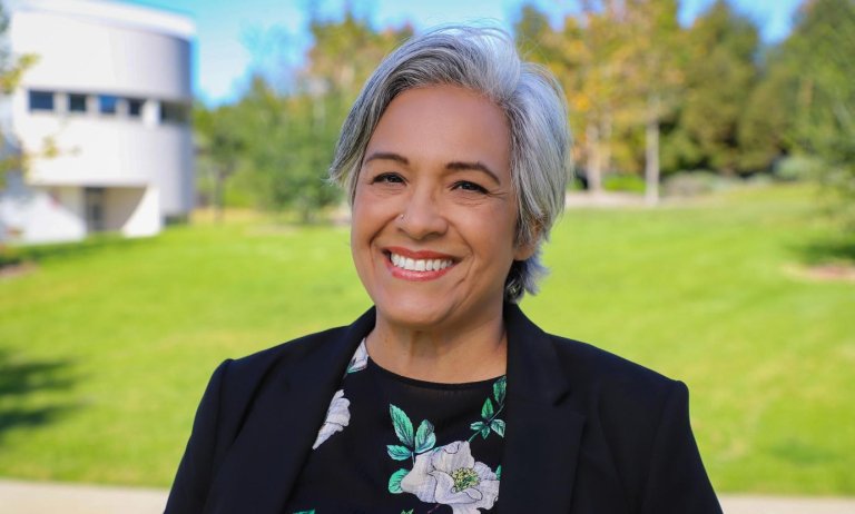 a woman with short white hair stands on Pitzer’s campus wearing a black blazer and top