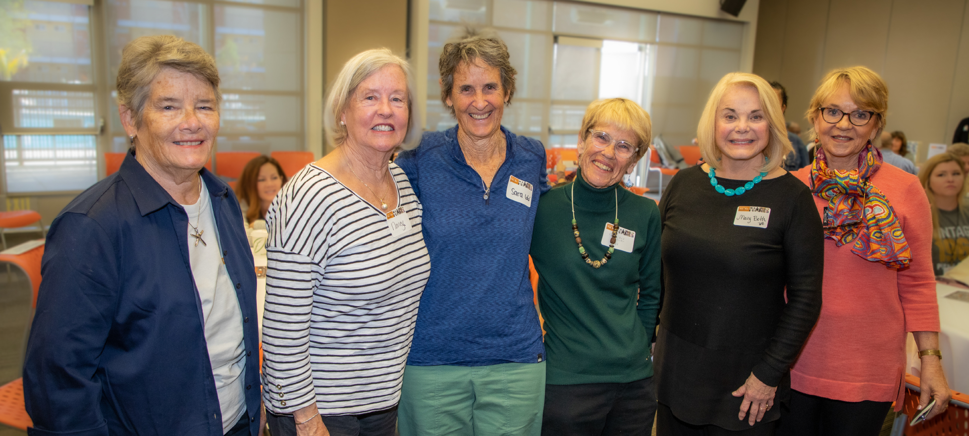 Six of Pitzer’s early alumnae stand with their arms around each other and smile. Behind them are people gathered in orange chairs around round tables.